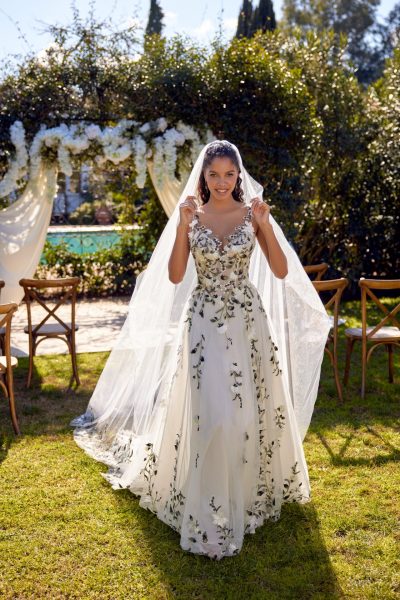 A woman standing in the grass wearing a wedding dress covered in embroidered flowers and a long veil (wedding dress trends)