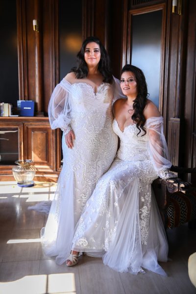 Two women wearing wedding dresses with sheer sleeves, one standing and one sitting. (wedding dress trends)