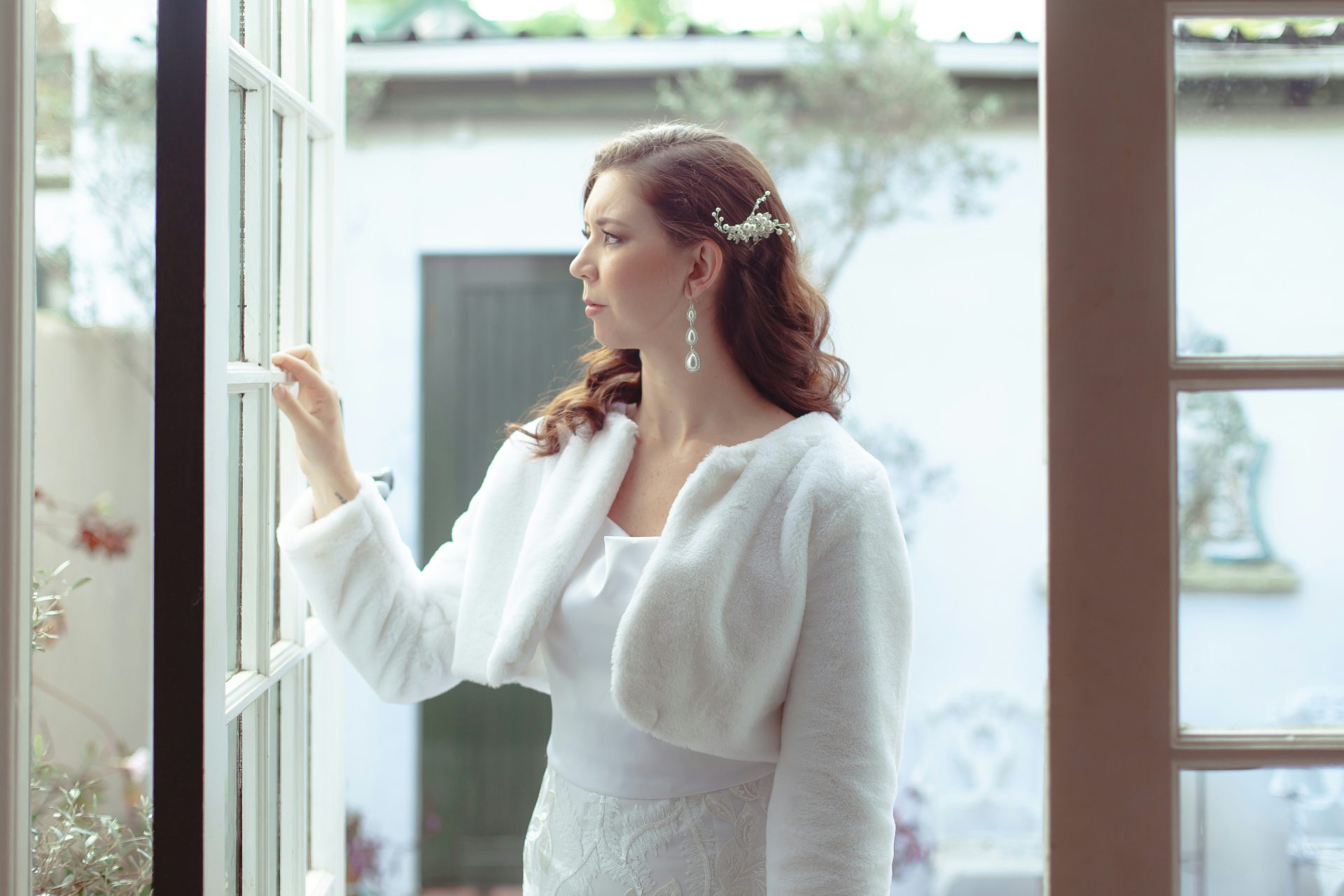 A bride, wearing a white fur jacket, looks out a window. (bridal accessories)