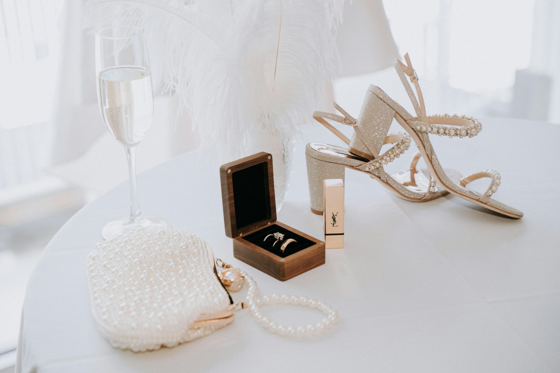 Pearl purse, a ring box, white shoes, and a glass of champagne on a white tablecloth. (bridal accessories)